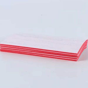 May include: A stack of white rectangular nail files with red edges. The files are stacked neatly, with the red edges creating a striped pattern. The files are on a white surface.