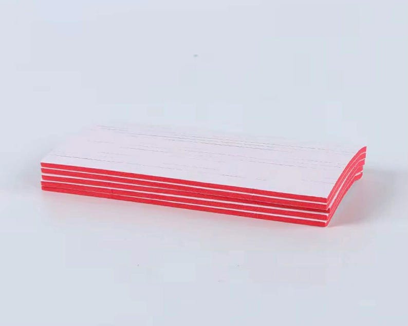 May include: A stack of white rectangular nail files with red edges. The files are stacked neatly, with the red edges creating a striped pattern. The files are on a white surface.