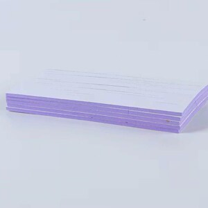 May include: A stack of rectangular, white paper sheets with lavender-colored edges. The paper is neatly stacked, with visible lines on the top surface. The background is a plain, light-colored surface.