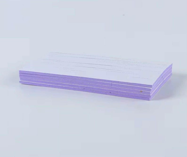 May include: A stack of rectangular, white paper sheets with lavender-colored edges. The paper is neatly stacked, with visible lines on the top surface. The background is a plain, light-colored surface.