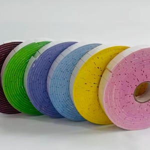 May include: A collection of colorful foam tape rolls in shades of burgundy, green, blue, yellow, and pink. Each roll is tightly wound, showing the textured surface of the foam. The rolls are arranged in a row, showcasing the variety of colors.