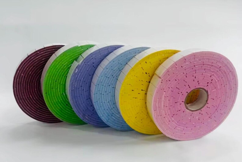 May include: A collection of colorful foam tape rolls in shades of burgundy, green, blue, yellow, and pink. Each roll is tightly wound, showing the textured surface of the foam. The rolls are arranged in a row, showcasing the variety of colors.