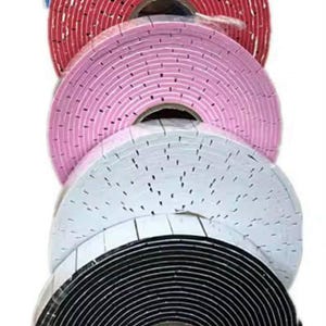 May include: A stack of rolls of foam tape in various colors: blue, red, pink, white, and black. The tape appears to be self-adhesive, with a grid pattern of small holes across the surface. The rolls are tightly wound and ready for use.