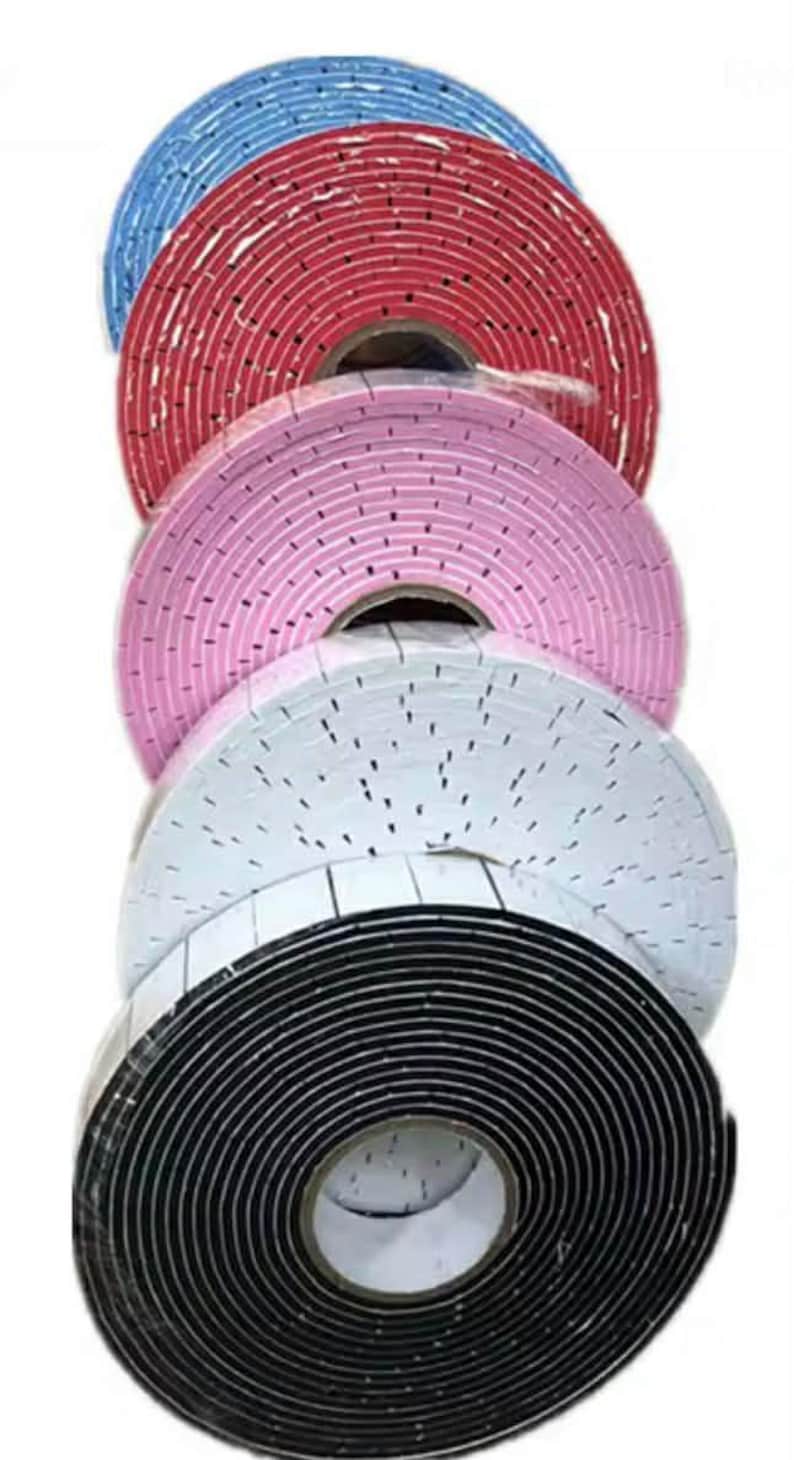 May include: A stack of rolls of foam tape in various colors: blue, red, pink, white, and black. The tape appears to be self-adhesive, with a grid pattern of small holes across the surface. The rolls are tightly wound and ready for use.