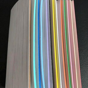 May include: A partially visible book with a white edge and colorful page edges. The page edges are in shades of blue, purple, yellow, orange, green, and pink. The book is on a dark surface.