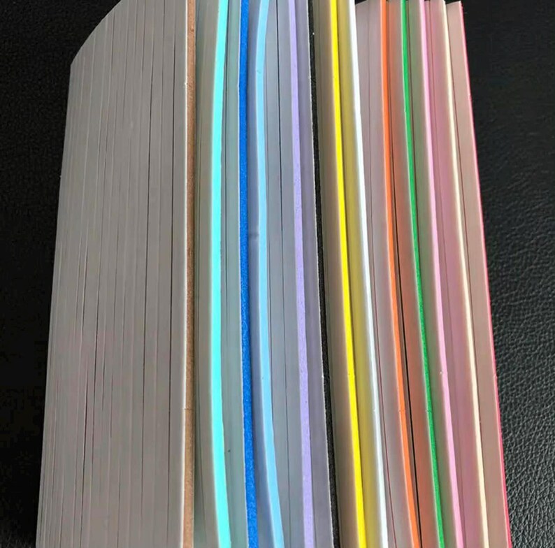 May include: A partially visible book with a white edge and colorful page edges. The page edges are in shades of blue, purple, yellow, orange, green, and pink. The book is on a dark surface.