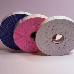 May include: Three rolls of foam tape in blue, pink, and gray. Each roll is circular with a white top layer and a colored foam core. The rolls are arranged in a row, with the blue roll on the left, the pink in the middle, and the gray on the right.