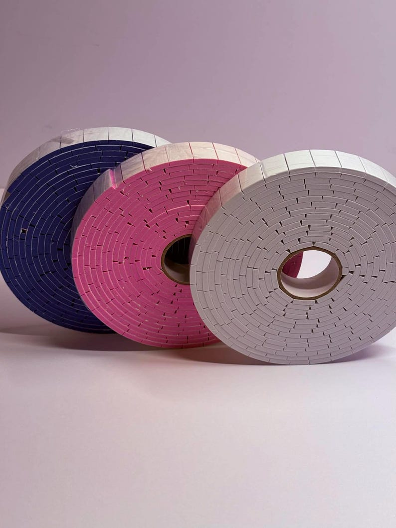 May include: Three rolls of foam tape in blue, pink, and gray. Each roll is circular with a white top layer and a colored foam core. The rolls are arranged in a row, with the blue roll on the left, the pink in the middle, and the gray on the right.
