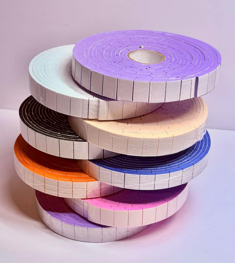May include: A stack of colorful foam tape rolls in various shades, including purple, white, black, orange, beige, blue, pink, and lavender. Each roll is neatly wrapped with a white backing, creating a visually appealing display of craft supplies.