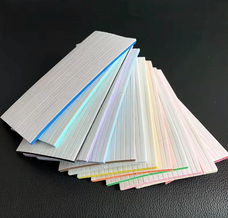 May include: A fan of colorful, textured, rectangular strips with various colored edges. The strips are arranged in a fanned-out pattern, showcasing a range of pastel and bright colors, including blue, green, pink, and yellow.