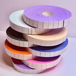 May include: A stack of colorful foam tape rolls in various shades, including purple, orange, blue, and pink. Each roll is neatly wrapped and has a grid pattern along the edges. The rolls are stacked on top of each other, creating a visually appealing display.
