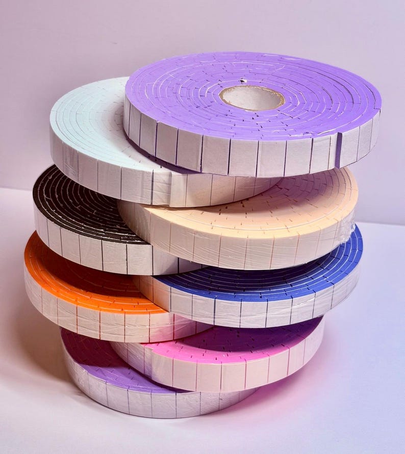May include: A stack of colorful foam tape rolls in various shades, including purple, orange, blue, and pink. Each roll is neatly wrapped and has a grid pattern along the edges. The rolls are stacked on top of each other, creating a visually appealing display.