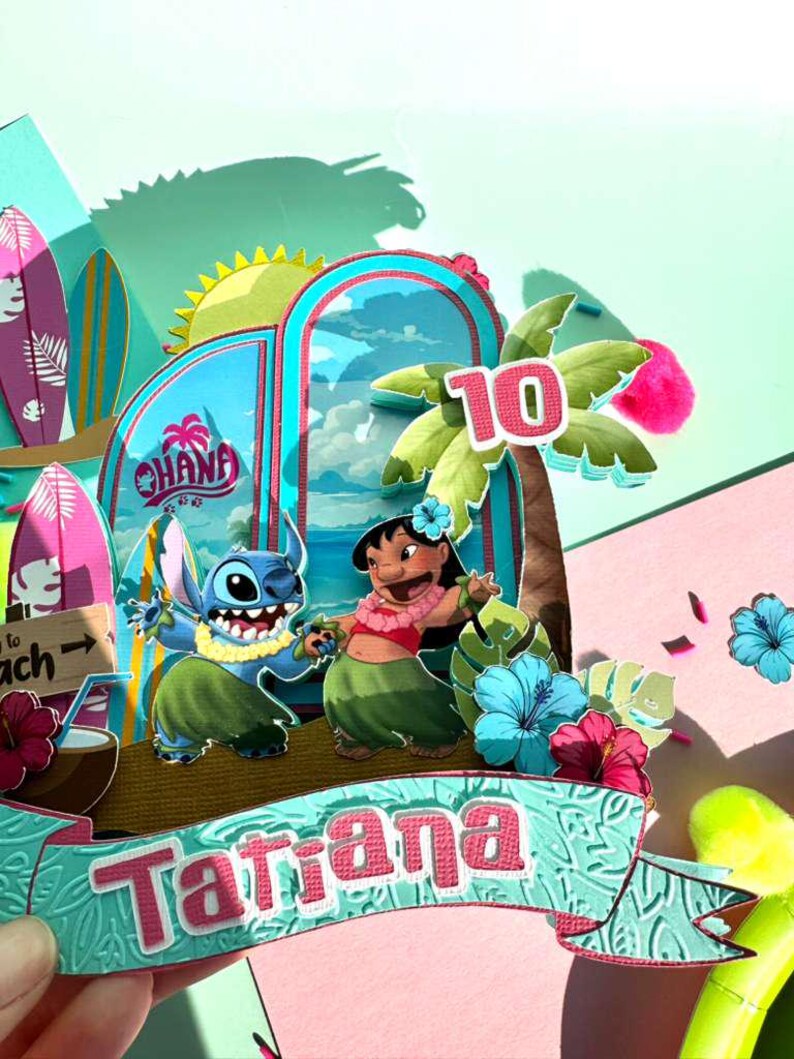 Puede incluir: Una colorida manualidad de papel con los personajes Stitch y Lilo de la pel&iacute;cula Lilo & Stitch. El dise&ntilde;o incluye tablas de surf, palmeras y la palabra "Ohana". El nombre "Tatiana" se muestra en una pancarta, con el n&uacute;mero "10".