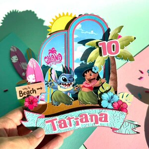 Puede incluir: Manualidad de papel colorida con personajes de la pel&iacute;cula Lilo & Stitch. El dise&ntilde;o incluye un arco azul con la palabra "Ohana", una palmera y el n&uacute;mero "10". Una pancarta dice "Tatiana".