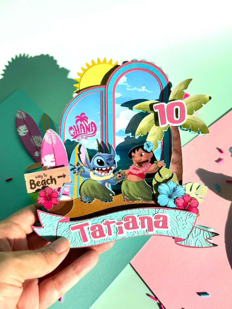 Puede incluir: Manualidad de papel colorida con personajes de la pel&iacute;cula Lilo & Stitch. El dise&ntilde;o incluye un arco azul con la palabra "Ohana", una palmera y el n&uacute;mero "10". Una pancarta dice "Tatiana".