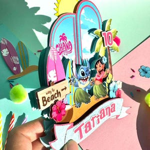 Puede incluir: Un adorno para tarta de cumplea&ntilde;os colorido y en capas con los personajes Stitch y Lilo de la pel&iacute;cula Lilo & Stitch. El dise&ntilde;o incluye una pancarta con el nombre "Tatiana", un letrero de "Way to Beach" y el n&uacute;mero "10".