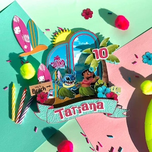 Puede incluir: Un adorno para tarta colorido con tem&aacute;tica tropical que presenta a los personajes Stitch y Lilo. El dise&ntilde;o incluye tablas de surf, palmeras y el nombre "Tatiana" con el n&uacute;mero 10. El fondo es una mezcla de rosa, verde y azul.