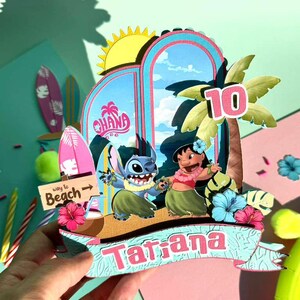 Puede incluir: Un adorno de pastel de cumplea&ntilde;os colorido con los personajes Stitch y Lilo de la pel&iacute;cula Lilo & Stitch. El adorno incluye una palmera, el n&uacute;mero 10 y el nombre "Tatiana" en una pancarta. El fondo tiene un tema de playa con tablas de surf y la palabra "Ohana".