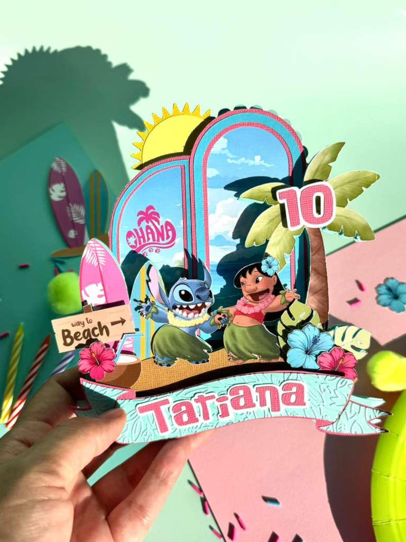 Puede incluir: Un adorno de pastel de cumplea&ntilde;os colorido con los personajes Stitch y Lilo de la pel&iacute;cula Lilo & Stitch. El adorno incluye una palmera, el n&uacute;mero 10 y el nombre "Tatiana" en una pancarta. El fondo tiene un tema de playa con tablas de surf y la palabra "Ohana".