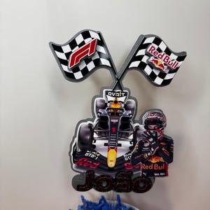 May include: A Red Bull Racing Formula 1 cake topper featuring a race car, driver, and checkered flags. The flags display the F1 logo and Red Bull branding. The name "Joao" is also included.