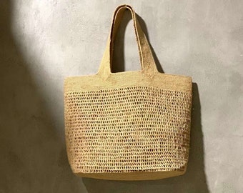 May include: A beige woven tote bag with a rectangular shape and two handles. The bag features an open weave design and is suspended against a grey wall. The bag's shadow is visible on the wall. The bag is made of natural materials.