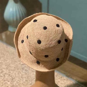 Może przedstawiać: Beżowy kapelusz przeciwsłoneczny z szerokim rondem i czarnymi kropkami. Kapelusz jest prezentowany na beżowej głowie manekina. Kapelusz jest wykonany z tkanego materiału.