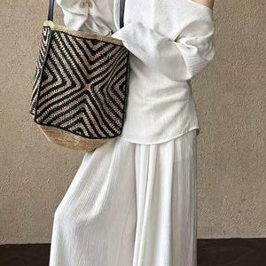 May include: A white off-the-shoulder top and wide-leg trousers paired with a black and beige geometric pattern tote bag. The tote bag has black leather straps. The outfit is a neutral colour palette.