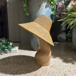 Peut inclure: Un chapeau en paille beige doté d'un bord large légèrement asymétrique et d'une calotte arrondie. Le chapeau est présenté sur un mannequin tête et cou de couleur beige lisse, mettant en valeur la texture tissée de la paille naturelle.