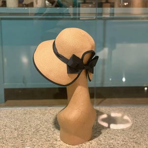 May include: A beige sun hat with a wide brim and a black ribbon. The hat has a black trim along the edge of the brim and a large black bow at the back. The hat is displayed on a mannequin head.