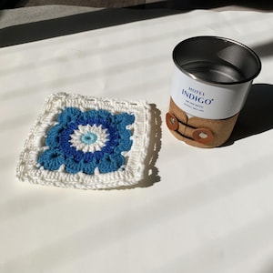 May include: A white crocheted square with a blue and white eye design in the center. The square is next to a white metal cup with a brown leather band and the text "Hotel Indigo" printed on the side.