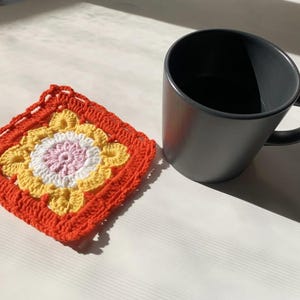 May include: A handmade, square crochet coaster with a floral design in shades of orange, yellow, white, and pink. A dark gray mug with a handle sits next to the coaster. The coaster is a decorative item.