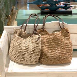May include: Two woven tote bags in shades of tan and brown with brown leather handles. The bags are made of a loosely woven material, creating an open, textured design. They are displayed on a white bench.