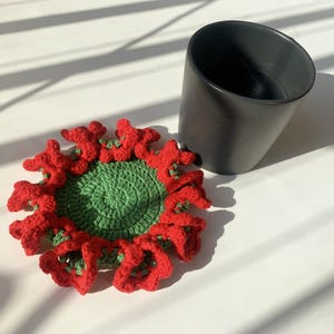 May include: A crocheted coaster with a green center and red flower-shaped edges. The coaster is sitting on a white surface next to a black mug.