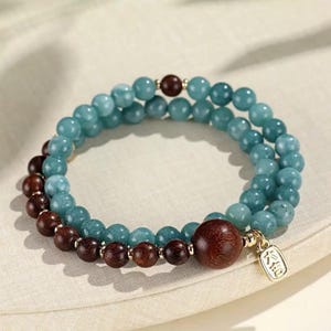 May include: Two-strand bracelet featuring light blue and brown beads. The bracelet has a mix of small and large round beads. A large wooden bead and a gold-colored charm with Chinese characters are also present.