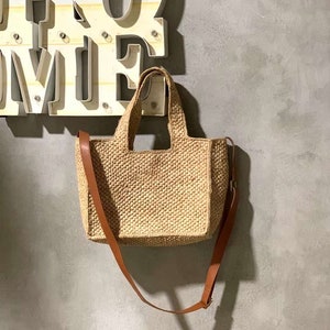 Könnte beinhalten: Eine geflochtene Strohtasche mit einem braunen Lederriemen und einem oberen Henkel. Die Tasche hängt an einer Wand mit einem Schild, auf dem "Home" steht.