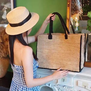 May include: A natural straw tote bag with black trim and handles. The bag is rectangular and appears to be made of woven straw. A straw hat with a black band is also visible.