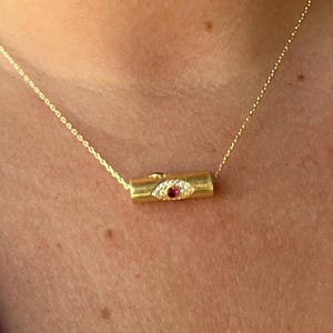 May include: A gold necklace with a cylindrical pendant. The pendant features an eye design with a central red gemstone and surrounding clear gems. The necklace is suspended from a fine gold chain.