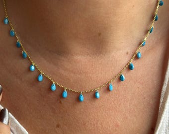 May include: A delicate gold necklace featuring small, teardrop-shaped turquoise beads. The necklace is displayed on a person's neck, showcasing the fine chain and the vibrant blue color of the beads. The necklace is a piece of jewelry.