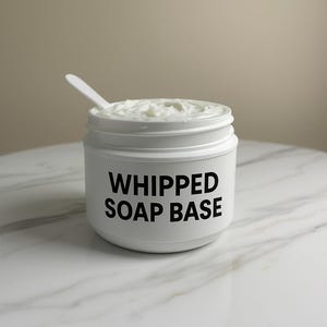 May include: A white jar labelled "WHIPPED SOAP BASE" contains a fluffy, white substance, with a small white spoon inside. The jar is on a marble surface, against a neutral background.