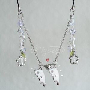 May include: A silver chain phone charm with two white cat charms, a heart charm, and a flower charm with green leaves. The chain also has white and iridescent beads.
