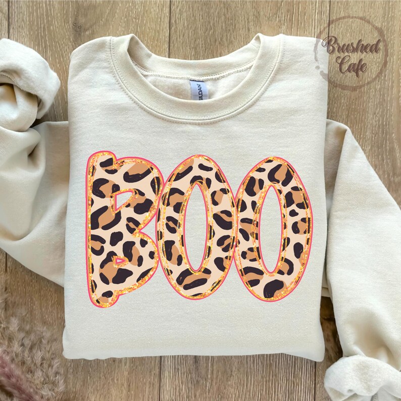May include: Cream-colored sweatshirt with the word "BOO" in large, leopard print letters. The letters have a pink outline and gold glitter accents. The sweatshirt is a crew neck style.