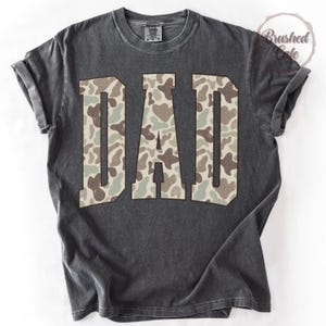 K&ouml;nnte beinhalten: Dunkelgraues T-Shirt mit dem Wort "DAD" in gro&szlig;en Buchstaben mit Camouflage-Muster. Das Shirt hat einen Vintage-Look mit abgenutztem Aussehen und hochgekrempelten &Auml;rmeln. Das T-Shirt hat einen Rundhalsausschnitt.