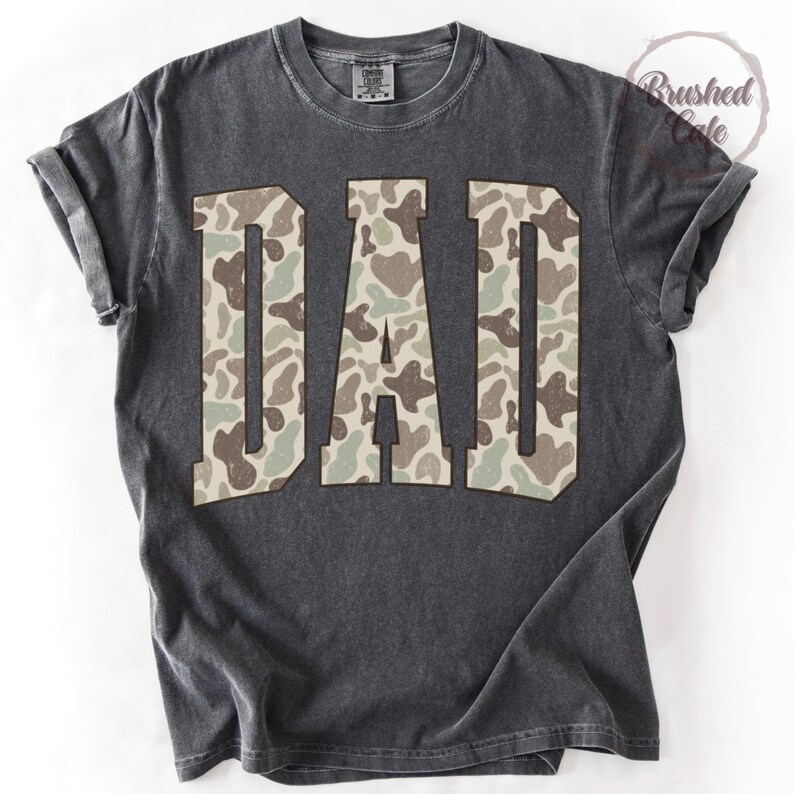 K&ouml;nnte beinhalten: Dunkelgraues T-Shirt mit dem Wort "DAD" in gro&szlig;en Buchstaben mit Camouflage-Muster. Das Shirt hat einen Vintage-Look mit abgenutztem Aussehen und hochgekrempelten &Auml;rmeln. Das T-Shirt hat einen Rundhalsausschnitt.