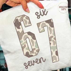May include: White pillow with the numbers "61" in a camouflage pattern, with the words "six" and "seven" in a script font. The pillow has a "Brushed Cafe" logo in the upper right corner.