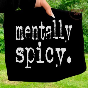 Puede incluir: Bolso tote negro con la frase "mentally spicy" en blanco, con una fuente audaz y desgastada. El bolso es sostenido por una mano con u&ntilde;as cuidadas, sobre un fondo de hierba verde y follaje.