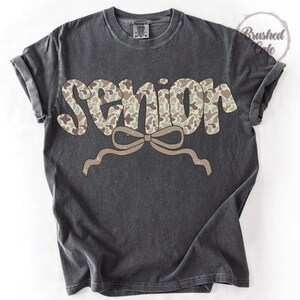 Pode incluir: Camiseta cinza escuro com a palavra "SENIOR" em um padr&atilde;o de camuflagem. Uma fita marrom clara est&aacute; abaixo da palavra. A camisa tem mangas enroladas e gola redonda.