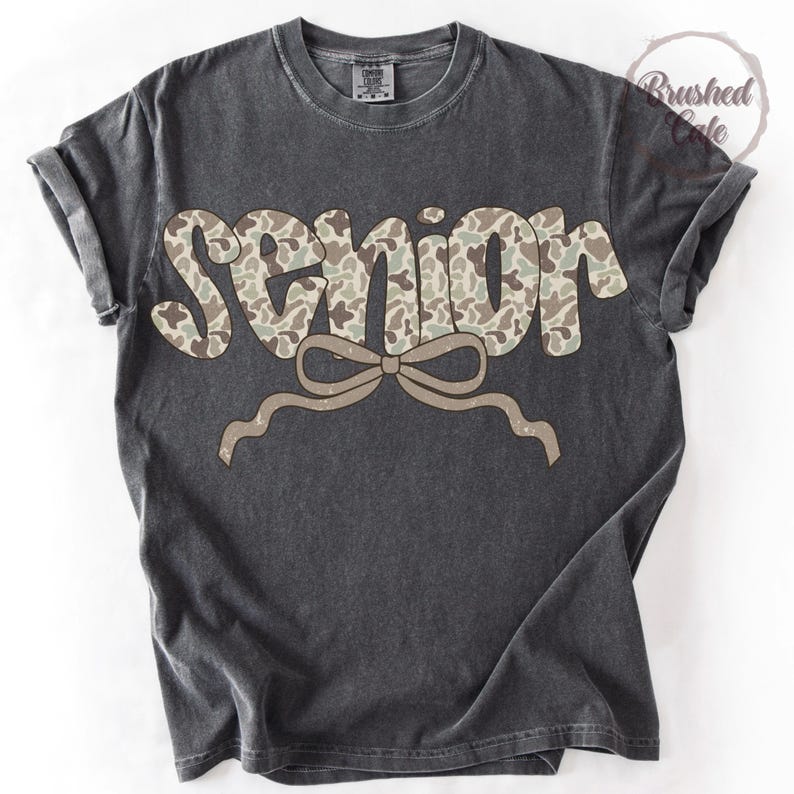 Pode incluir: Camiseta cinza escuro com a palavra "SENIOR" em um padr&atilde;o de camuflagem. Uma fita marrom clara est&aacute; abaixo da palavra. A camisa tem mangas enroladas e gola redonda.