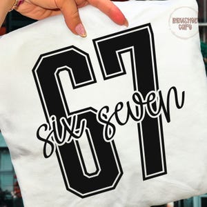 May include: White t-shirt with a large black number 67 design. The number is outlined in white and features the words "Six seven" in a cursive font. The shirt is held by a hand.