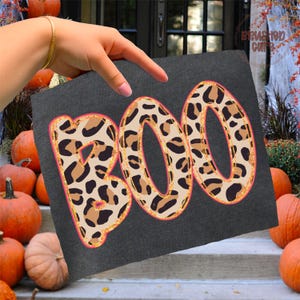 May include: A dark gray square with the word "BOO" in large, leopard print letters. The letters have a gold glitter outline and a pink border. Pumpkins are in the background, suggesting a Halloween theme.