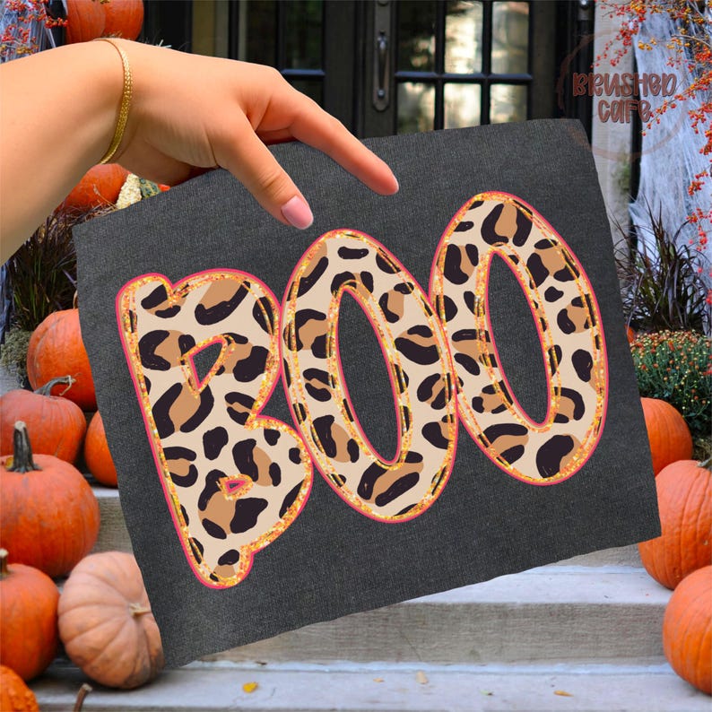 May include: A dark gray square with the word "BOO" in large, leopard print letters. The letters have a gold glitter outline and a pink border. Pumpkins are in the background, suggesting a Halloween theme.